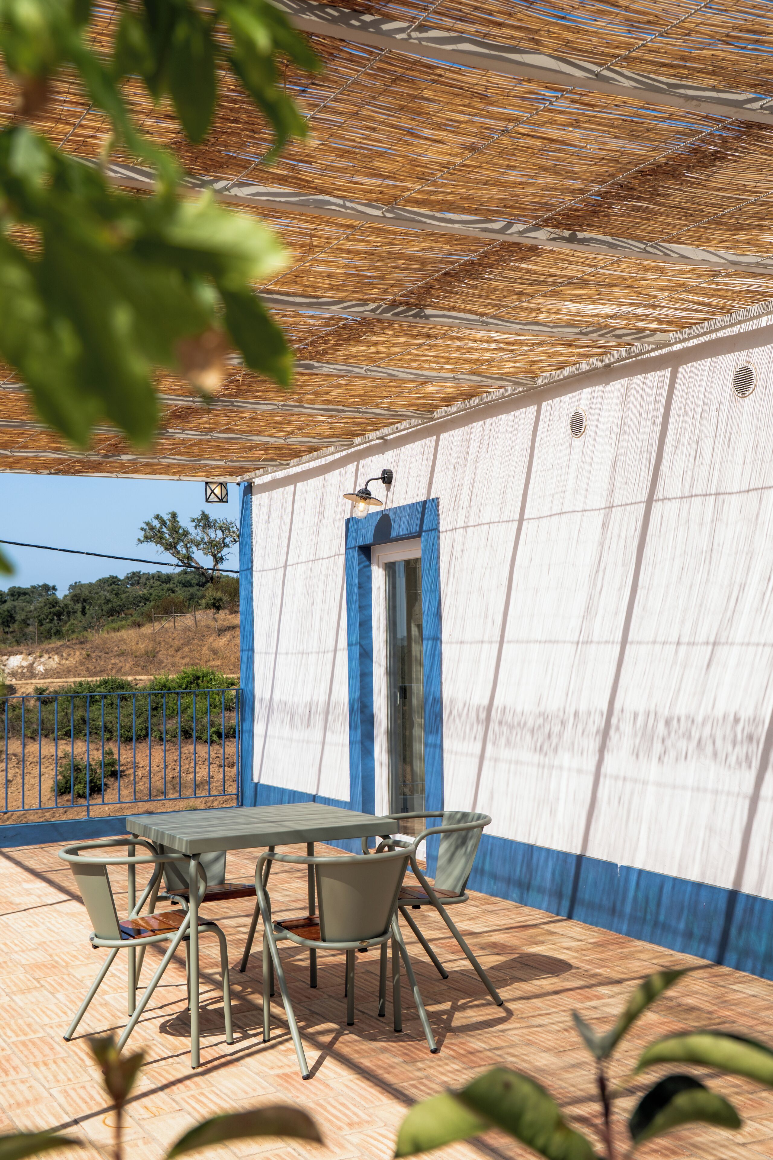 Un patio exterior con mesa y sillas, una puerta con marco azul y una pérgola.