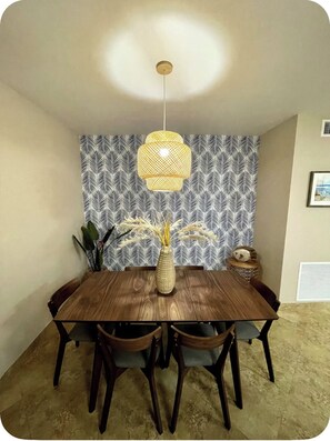A dining area with a wooden table, four chairs, a hanging light fixture, a patterned wallpaper, and a vase with dried plants.