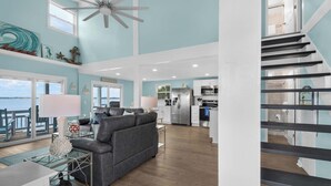 A spacious living room with a grey sofa, a glass coffee table, and a view of the sea through large windows. - Sandy Sunset Sound front 4BR Home Two Decks w/ Sunset Views Boat Dock (Navarre)
