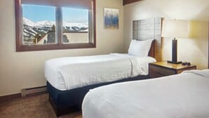 Una camera d'albergo con due letti, una finestra vista montagne, un comodino con una lampada e un quadro appeso al muro.