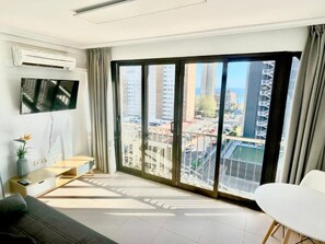 A modern living room with a large sliding glass door to a city view, a flat-screen TV mounted on the wall, and a minimalist furniture setup.