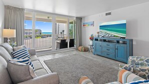 A living room with a sofa, a television, and a view of the beach through large windows. - Shoreline Towers 3033 (Destin)