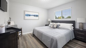 Een slaapkamer met een bed, nachtkastjes, een bureau, een stoel, een raam en een schilderij aan de muur.