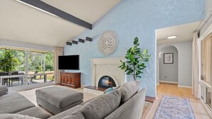 A spacious living room with grey sofas, a flat-screen TV, a fireplace, and a large window overlooking a garden. - Quiet Riverside Retreat 3bd/2ba (Auburn)