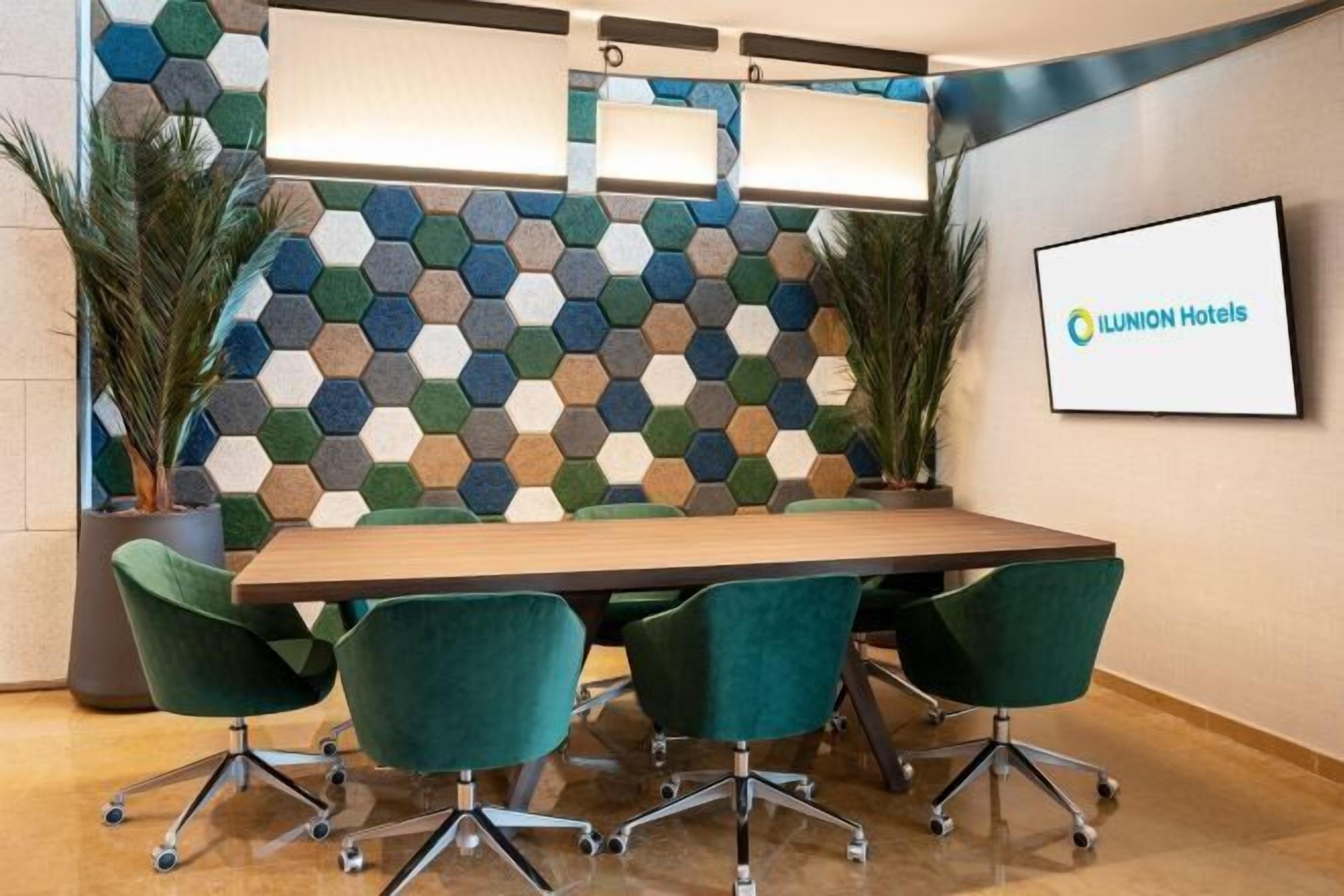 Un salón de conferencias con una mesa de madera, sillas verdes, un revestimiento de pared con patrón hexagonal y un cartel de ILUNION Hotels.