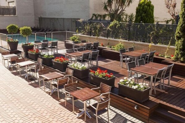 An outdoor patio with multiple tables and chairs, surrounded by potted plants and a swimming pool. - Ilunion Valencia 4 (Valencia)