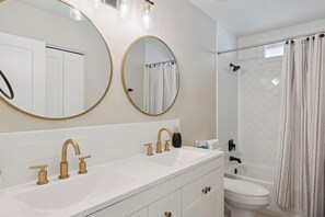 Een badkamer met twee wastafels, gouden kranen, een toilet en een douche met een witte backsplash van visgraatmozaïek.
