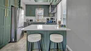 Une cuisine avec des meubles de rangement verts, un réfrigérateur en acier inoxydable, un îlot central blanc avec deux tabourets et une fenêtre avec des stores.