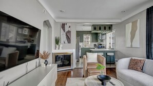 Un salon moderne avec un canapé, une table basse et une cheminée. On aperçoit au fond une cuisine avec des meubles verts et un coin repas.