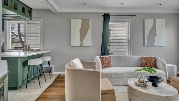 Un salon moderne avec un canapé, un pouf et une table basse, un coin cuisine avec des tabourets de bar et des œuvres d’art accrochées aux murs.