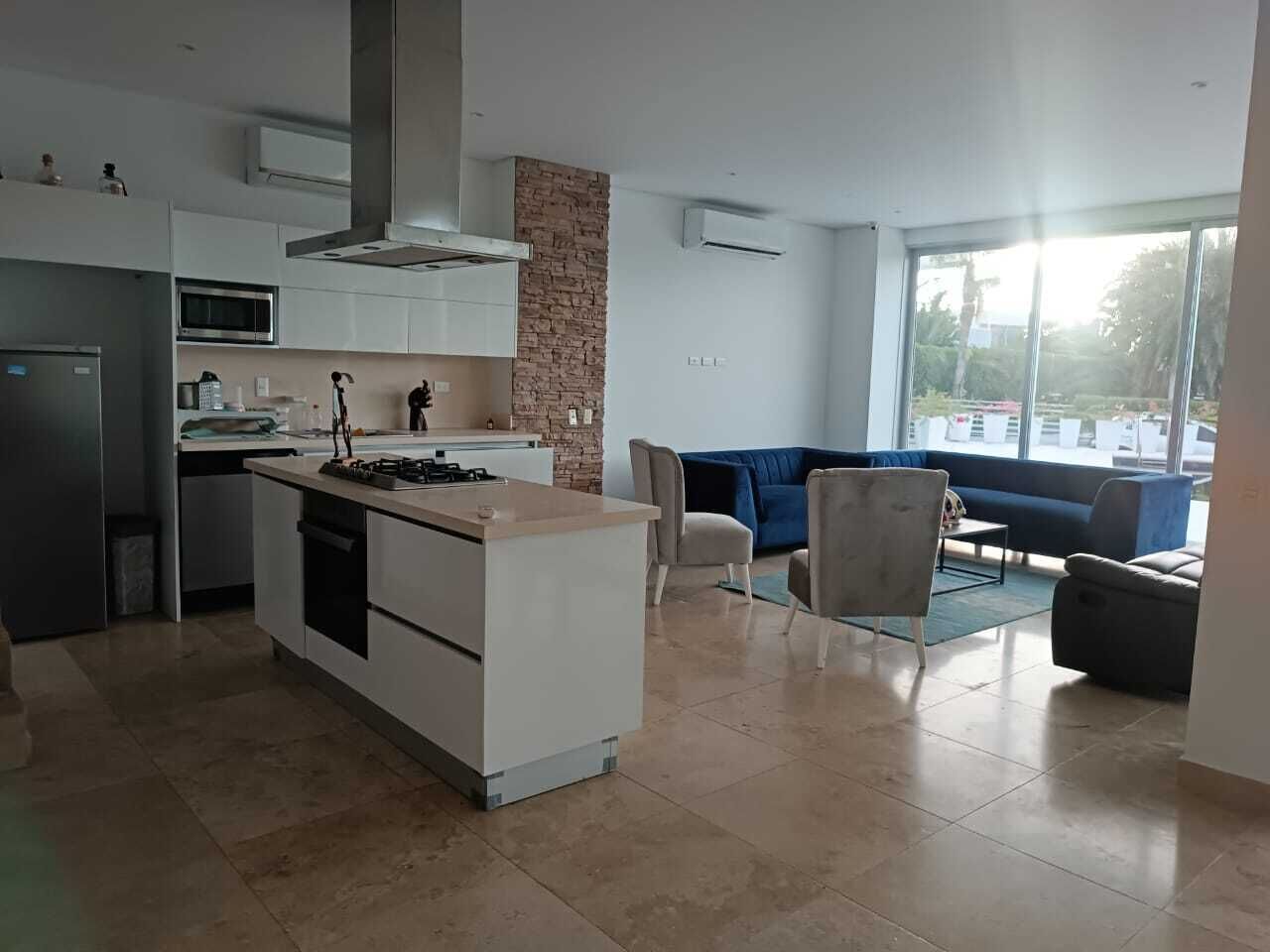 A modern kitchen with a central island, white cabinetry, and a stone accent wall. There is a dining area with a blue sofa and a small table.