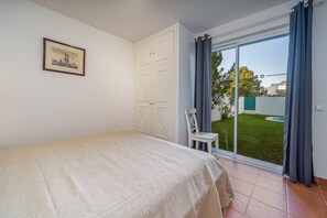 A bedroom with a bed, a white wardrobe, a chair, and a sliding glass door leading to a garden.