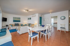 A living room with a sofa, TV, dining table, chairs, and a kitchen in the background.