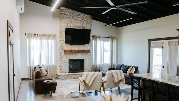 A spacious living room with a stone fireplace, a flat-screen TV, a dining area with a marble table, and a bar area. - Freedom Quarters

~Farm Setting~ (Fordland)