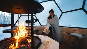 Eine Person, in eine Decke gehüllt, sitzt am Feuer in einer geodätischen Kuppel mit Blick auf schneebedeckte Bäume.