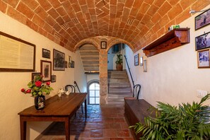 Un couloir aux murs de pierre, avec une table en bois, des chaises et un escalier menant à un niveau supérieur.