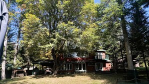 A red building with a balcony, surrounded by trees and a grassy area. - ADK Channel House - Spacious, waterfront home. Huge boathouse with top sun deck (Old Forge)