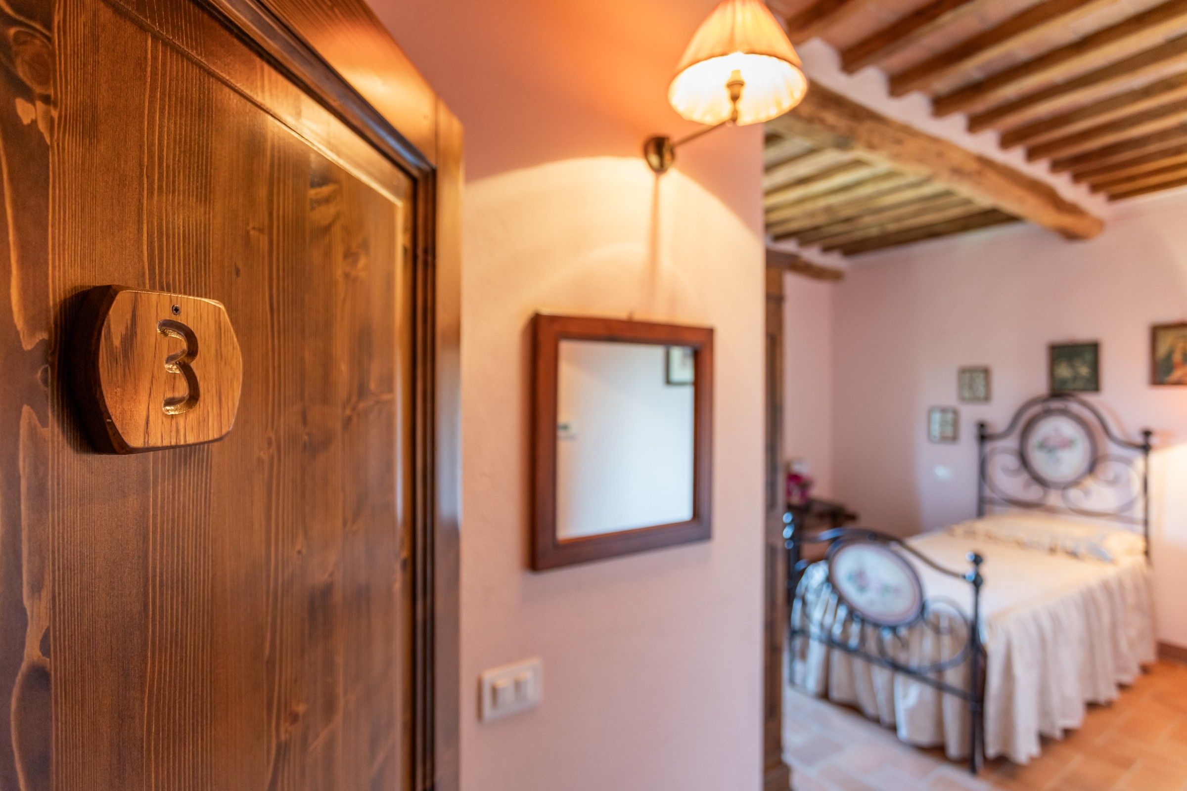 A wooden door with a number plate, a mirror on the wall, a bed with a metal frame, and a ceiling with wooden beams.