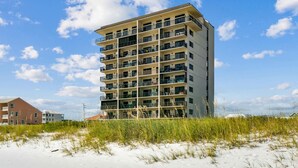 Un hotel de varios pisos con balcones, situado cerca de una playa de arena con poca vegetación.