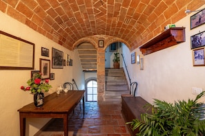 Un couloir aux murs de pierre, avec une table en bois, des chaises et un escalier menant à un niveau supérieur.