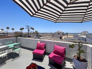 A rooftop terrace with a striped awning, two wicker chairs with pink cushions, a small table, and a potted plant. - Saylors Aweigh Guest Suite (Seal Beach)