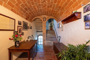 A stone-walled corridor with a wooden table, a vase of flowers, and a staircase leading to an upper level.