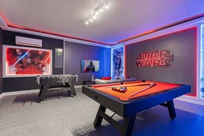 A room with a pool table, a foosball table, Star Wars-themed wall art, and a Star Wars logo.