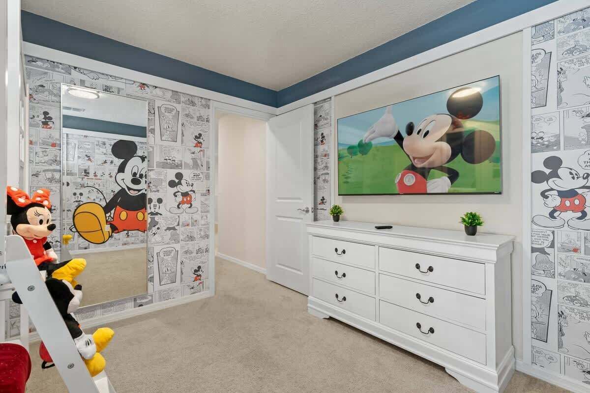 Una habitación con una gran televisión de pantalla plana montada en la pared, un aparador blanco con cajones y papel tapiz con motivos de Mickey Mouse.