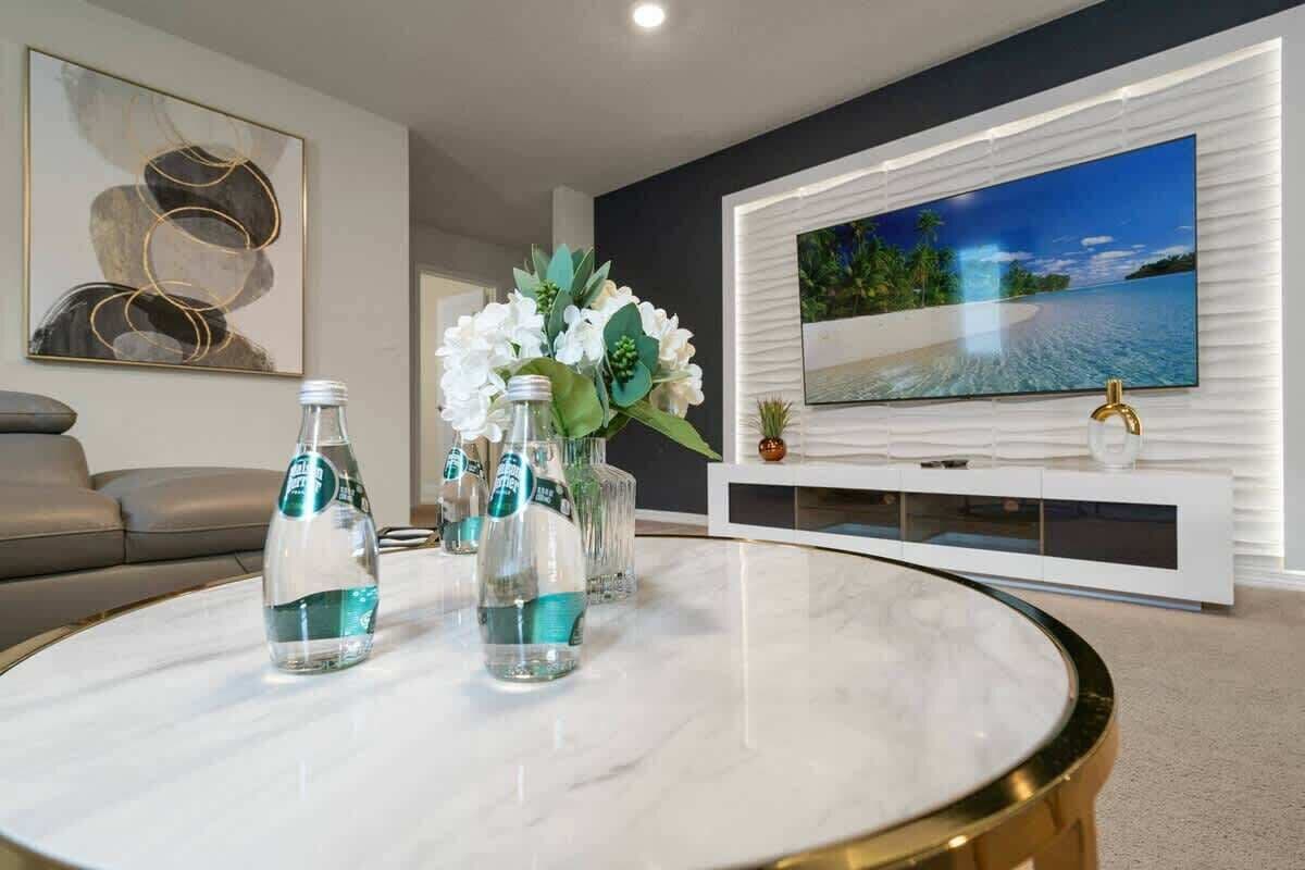 A living room with a round marble coffee table, a sofa, a TV mounted on a white wall, and a decorative plant arrangement.