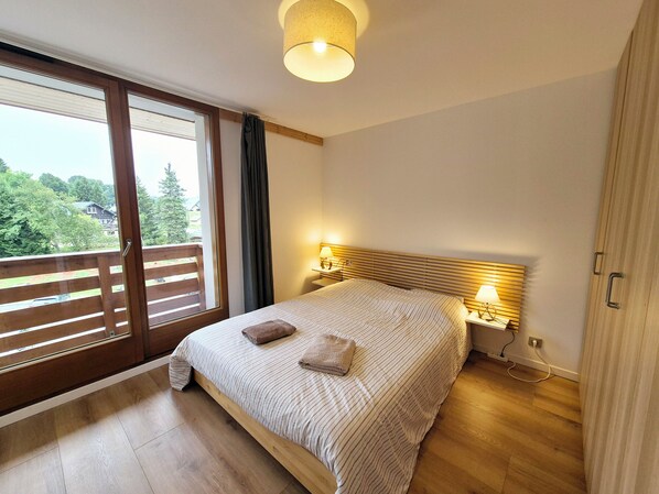 A bedroom with a large bed, wooden headboard, two bedside lamps, and a view of greenery through a sliding door.