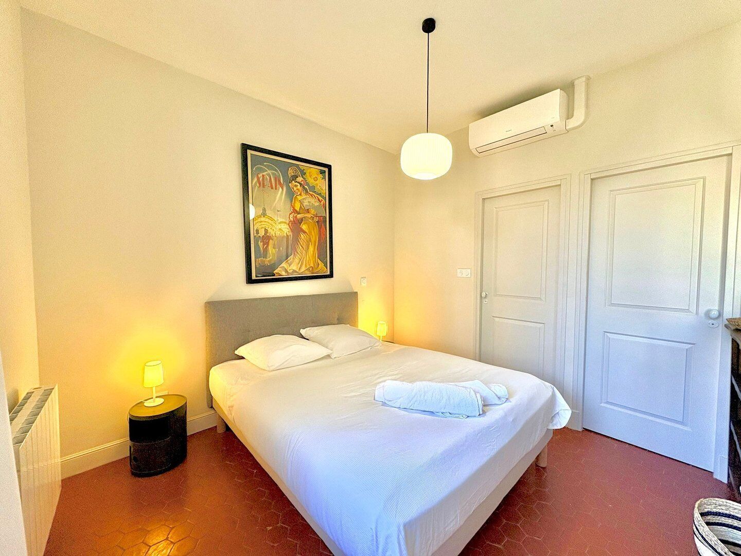 Una habitación de hotel con cama, un cuadro, una lámpara, aire acondicionado y una puerta.