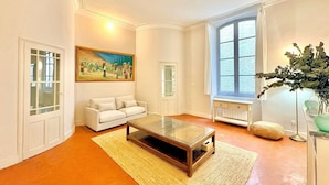 A living room with a glass coffee table, white sofa, a painting, a window with curtains, and a plant. - Hôtel Boudon 4, Luxury 1BR in Écusson (Nîmes)