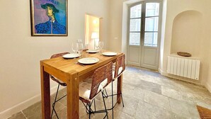A dining area with a wooden table set for four, a wicker chair, a framed painting, a door with glass panels, and a radiator. - Hôtel Boudon 4, Luxury 1BR in Écusson (Nîmes)