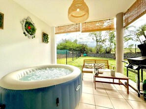 A hot tub in a sunlit room with a view of a garden, a table with chairs, and a grill outside. - Le Kerbo, Peace & Private Jacuzzi in Bras-Panon (Bras-Panon)