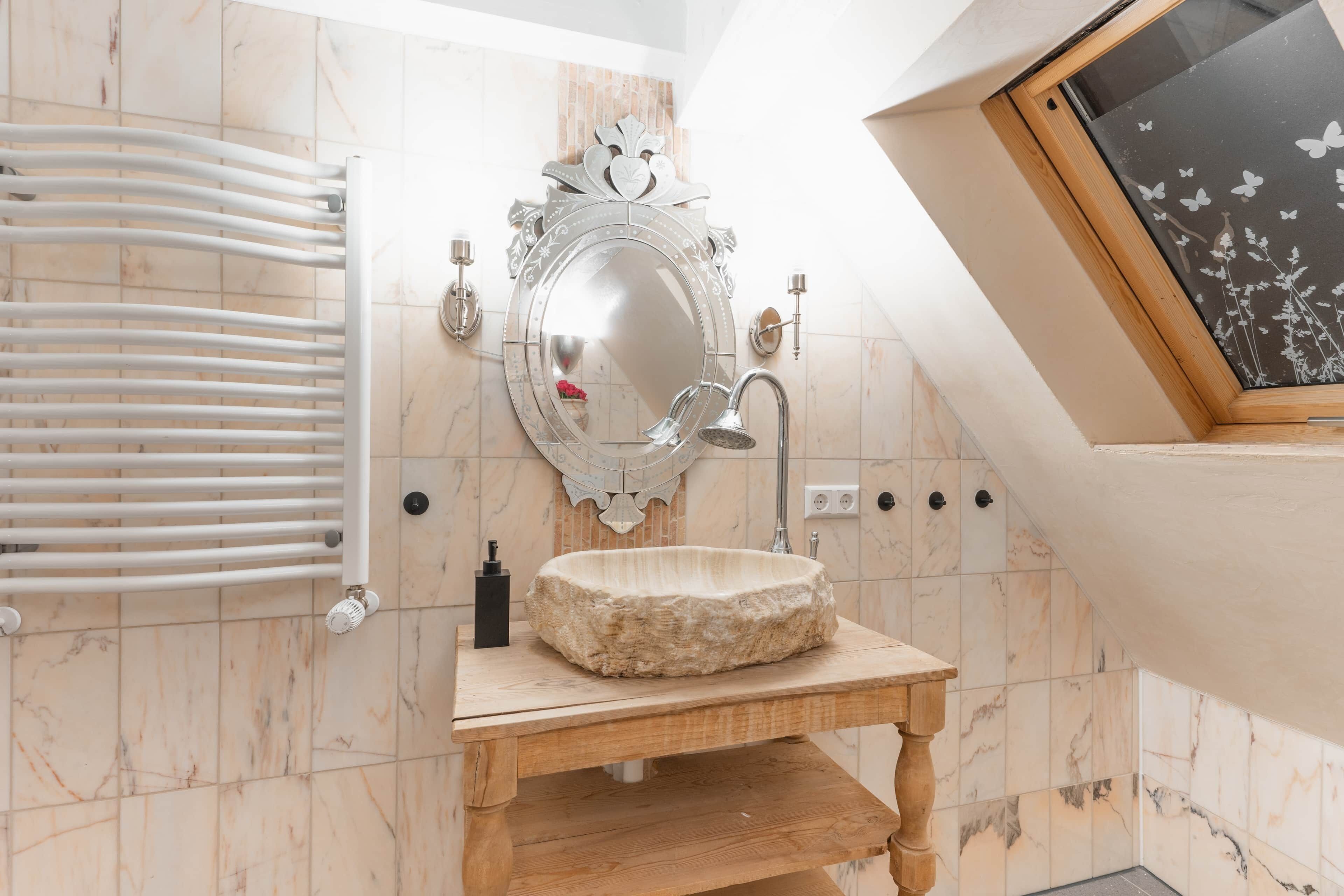 Un baño con pared de azulejos de mármol, radiador blanco, tocador de madera con lavamanos de piedra, espejo grande con marco decorativo y tragaluz con un adhesivo de mariposa.