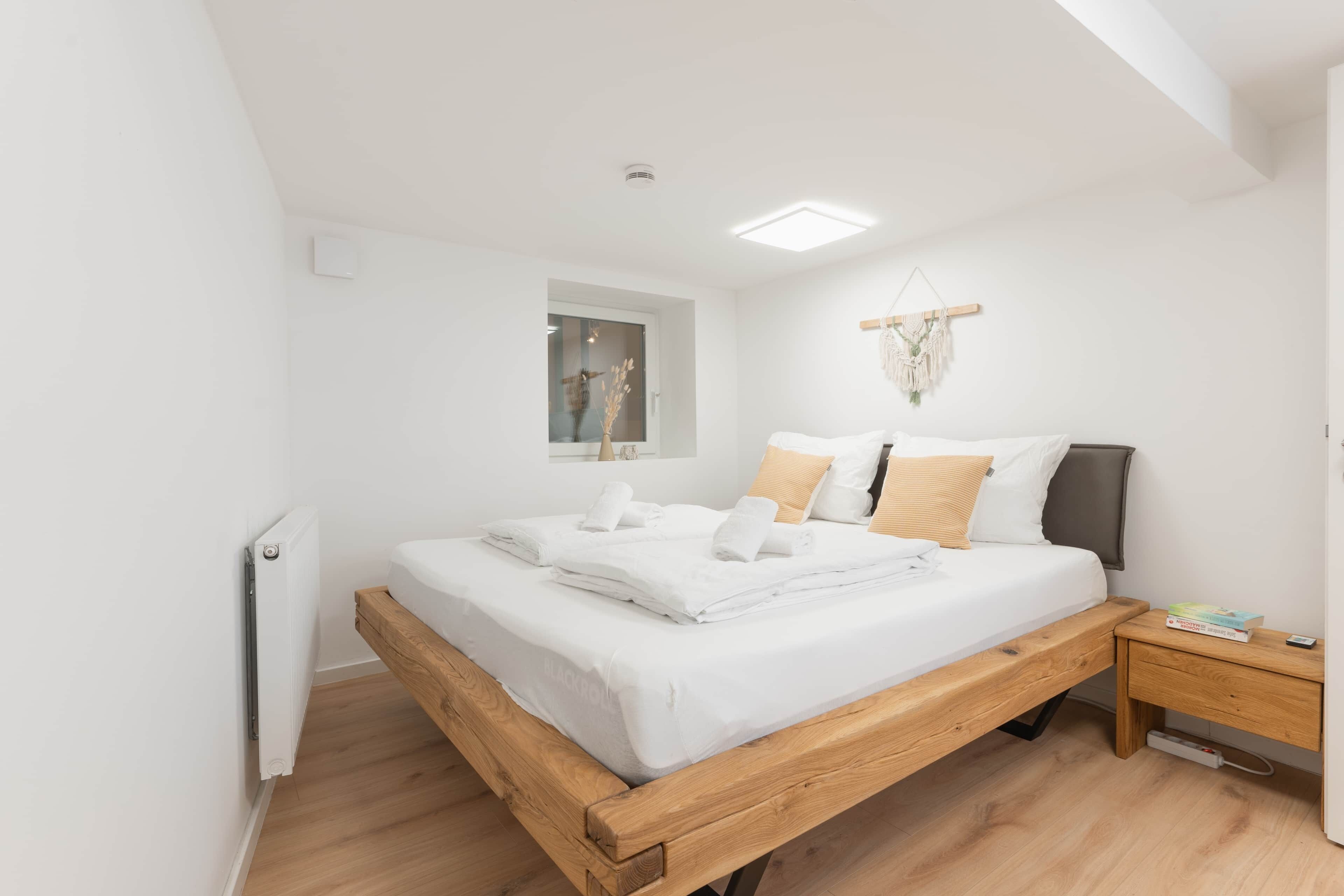 Un dormitorio moderno con una cama de madera, ropa de cama blanca, una mesita de noche, una pequeña lámpara de noche y una decoración de macramé en la pared.