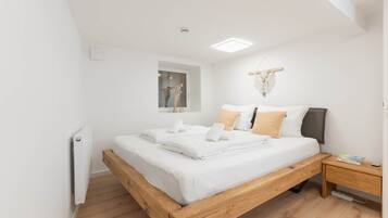 Une chambre moderne avec un lit en bois, du linge de lit blanc, une table de chevet, une petite lampe de chevet et une décoration en macramé fixée au mur.