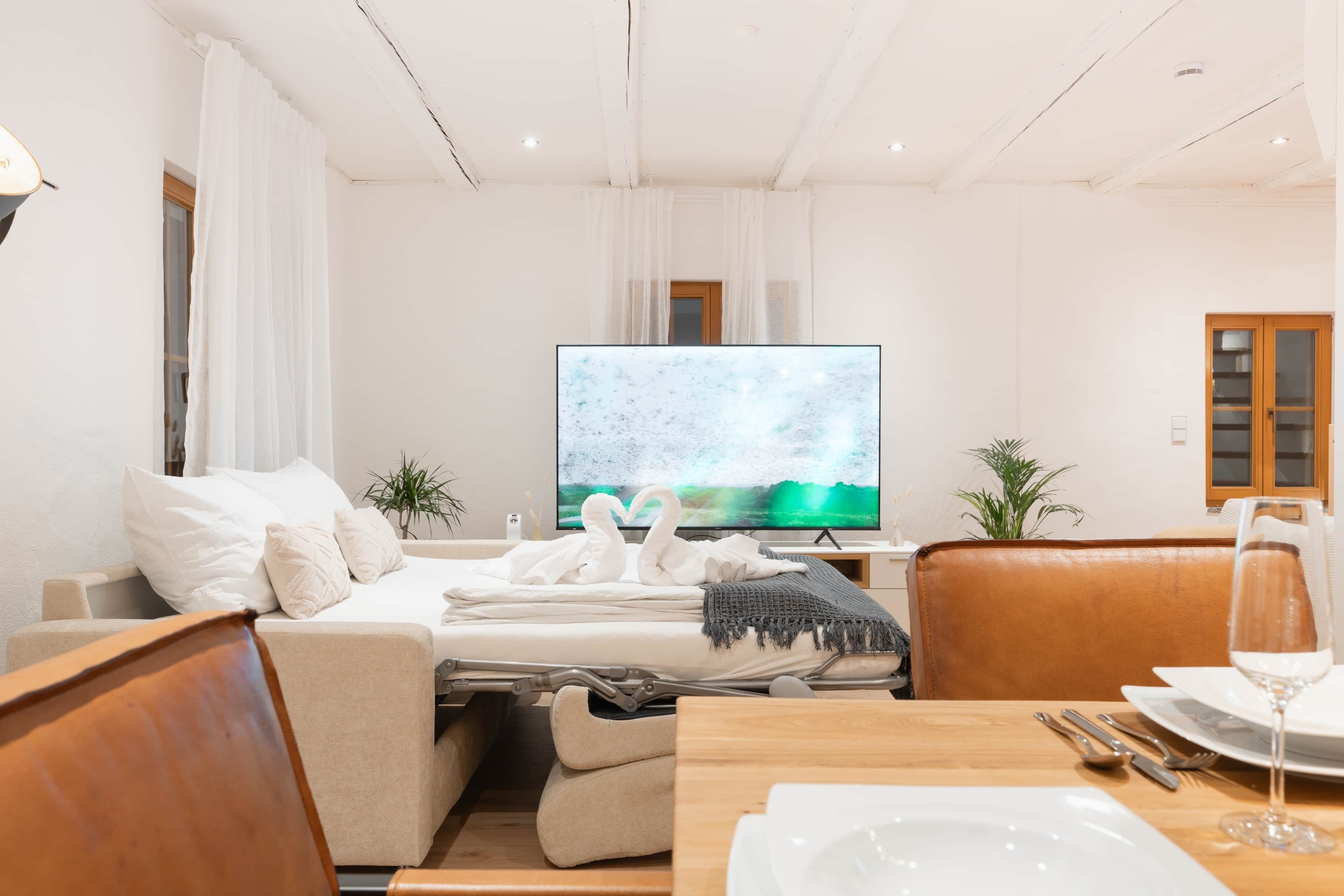 A modern hotel room with a sofa, a dining table set with a glass of wine, and a flat-screen TV displaying a nature scene.