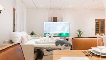 A modern hotel room with a sofa, a dining table set with a glass of wine, and a flat-screen TV displaying a nature scene.