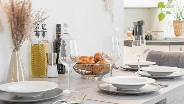 A dining table set with plates, glasses, and a bowl of croissants, with bottles and a vase in the background. - Untersee Suite | Lake nearby – Terrace - Workspace (Radolfzell)