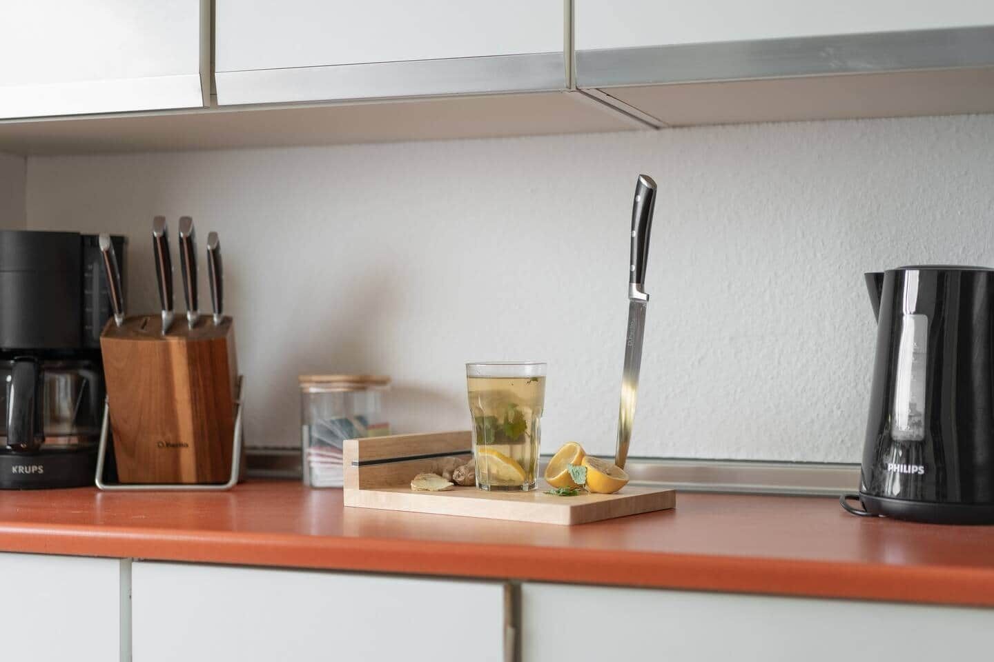 Una encimera de cocina con una cafetera, un cuchillo, un vaso de té y un bloque de madera con rodajas de limón.