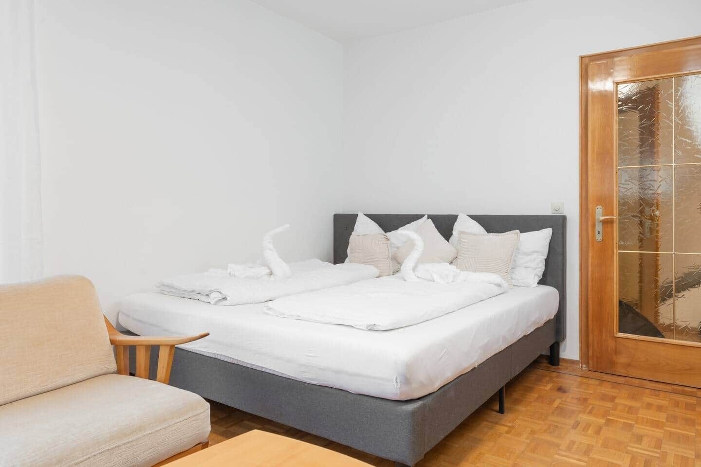 Un lit bien fait, avec du linge de lit blanc et des cygnes décoratifs, une tête de lit rembourrée de couleur grise, une porte en bois avec des panneaux de verre et un plancher en bois.