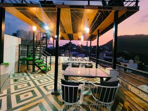 An outdoor terrace with tables and chairs, a staircase, and a view of the cityscape at dusk. - Hotel L S Restaurant & Spa (Udaipur)
