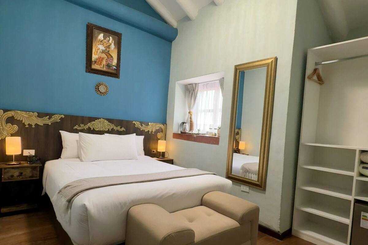 Ein Schlafzimmer mit Bett, Nachttischen, einer Bank, einem Kleiderschrank, einer Chandelierleuchtre und einem gerahmten Bild an der Wand.