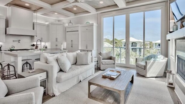 A modern kitchen with white cabinetry, a dining area with a white sofa and chairs, and large windows offering a view of a residential area. - Caspian Escape (Santa Rosa Beach)