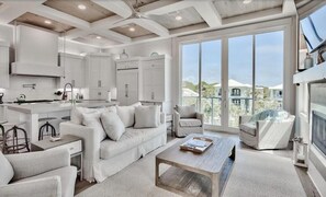 A modern kitchen with white cabinetry, a dining area with a white sofa and chairs, and large windows offering a view of a residential area. - Caspian Escape (Santa Rosa Beach)