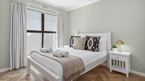 A neatly made bed with rolled towels, a bedside lamp, and a small plant on a white nightstand. - 17 Megan's Place - Two Bedroom Apartment, Sleeps 3 (Blouberg)