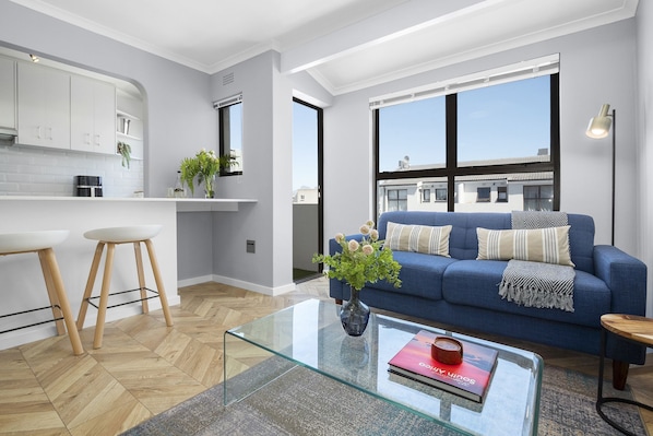 A modern apartment with a kitchen, a living area with a blue sofa, and large windows.