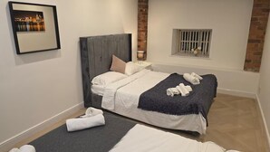 A hotel room with two beds, a framed picture on the wall, a brick accent wall, and a window with a view. - Liverpool Penthouse - Sleeps 8 - City Centre (Liverpool)