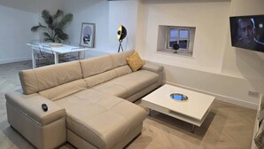 A modern living room with a beige sectional sofa, a white coffee table, a dining table with chairs, a floor lamp, and a TV mounted on the wall. - Liverpool Penthouse - Sleeps 8 - City Centre (Liverpool)